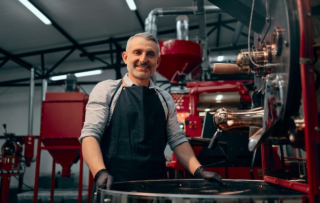 Retrato sorridente torrador de café em pé na máquina de torrefação e olhando para a câmera Foco seletivo