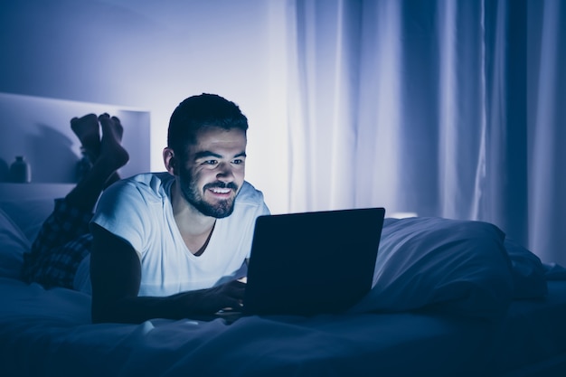 Retrato de su chico barbudo alegre alegre atractivo agradable acostado en la cama usando computadora portátil digital charlando pasar tiempo en la noche tarde en casa habitación oscura casa plana