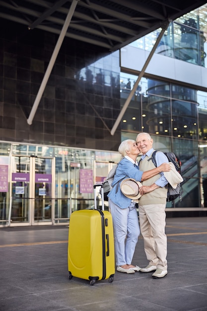 Retrato de tamaño completo de una mujer amorosa besando a un turista masculino contento sonriente antes de la terminal del aeropuerto
