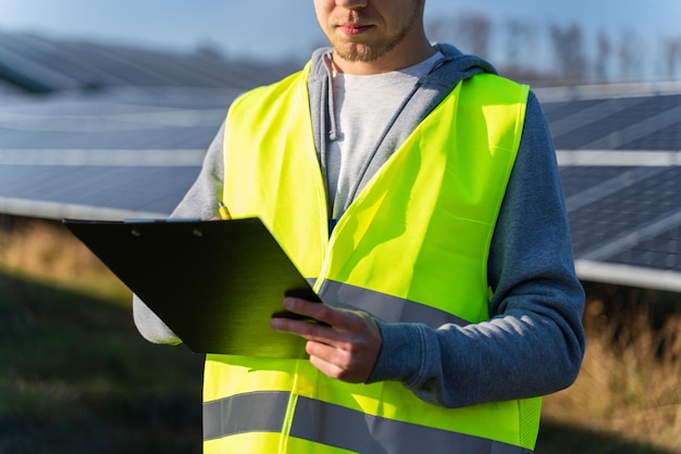 Retrato de tecnólogo masculino en uniforme con casco protector Hombre adulto sosteniendo un portapapeles en sus manos de pie en el campo con paneles solares