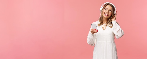 Retrato de una tierna y hermosa chica caucásica rubia vestida de blanco soñando despierta mientras escucha la pista romántica favorita usar auriculares sostener el teléfono móvil mirar hacia arriba pensativo disfrutar de la pista favorita