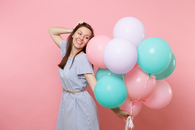 Retrato de tierna mujer joven sonriente en vestido azul sosteniendo coloridos globos de aire manteniendo la mano cerca de la cabeza aislada sobre fondo rosa brillante. Fiesta de cumpleaños, concepto de emociones sinceras de personas.