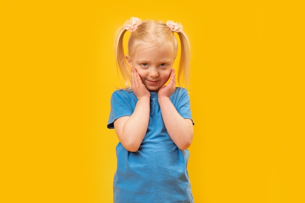 Retrato de una tímida niña preescolar con dos colas de caballo Niña rubia de 45 años en camiseta azul sobre fondo amarillo