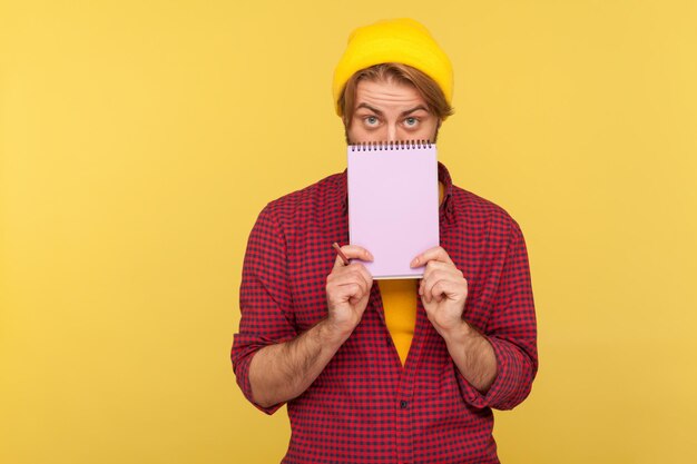 Retrato de un tipo barbudo hipster con sombrero de beanie y camisa a cuadros de pie cubriendo la mitad de la cara con un cuaderno de papel, mirando a la cámara. Disparo de estudio interior aislado sobre fondo amarillo.