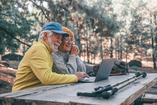 Retrato de un tiro en la cabeza cerca de una linda pareja de personas de mediana edad que usan computadoras al aire libre sentadas en una mesa de madera en el bosque de la montaña en la naturaleza con árboles a su alrededor