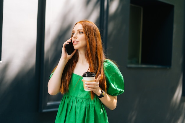 Retrato de tiro medio de mujer joven atractiva en vestido verde sosteniendo una taza de café para llevar, hablando por teléfono móvil caminando en la calle de la ciudad en un día soleado de verano, mirando a otro lado.