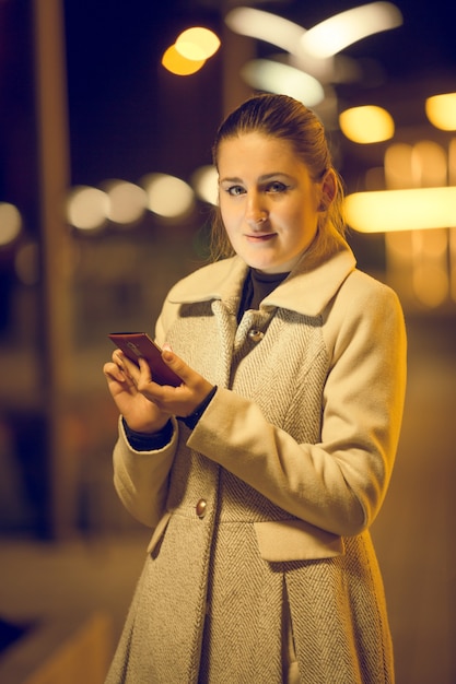 Retrato tonificado à noite de uma jovem elegante usando o telefone celular na rua