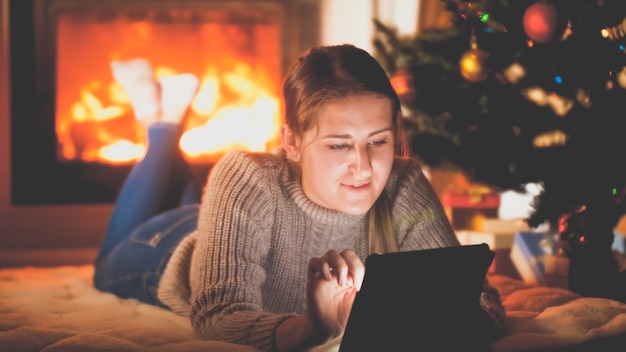 Retrato tonificado de uma mulher sorridente com um tablet deitado no chão ao lado do firepalce e da árvore de Natal