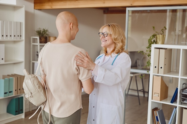 Retrato en tonos cálidos de la alegre doctora saludando al paciente calvo y felicitándola por la recuperación del cáncer, espacio de copia