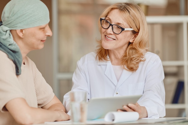 Retrato en tonos cálidos de doctora sonriente hablando con una mujer calva y mostrando datos en tableta digital durante la consulta sobre alopecia y recuperación del cáncer, espacio de copia