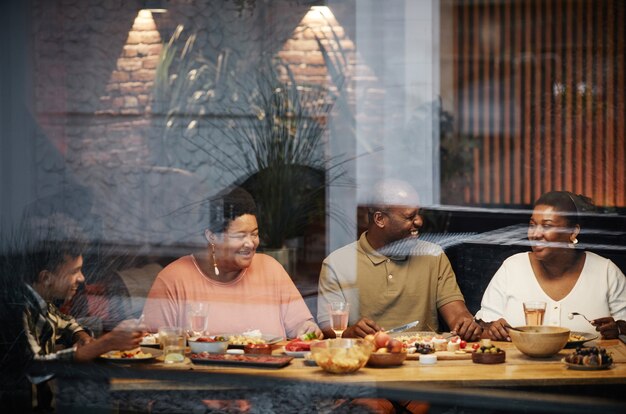 Retrato de tonos cálidos de familia afroamericana feliz disfrutando de la cena juntos en la terraza detrás de un vidrio ...