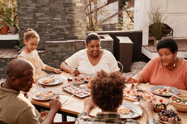 Retrato de tonos cálidos de la familia afroamericana feliz sentado en la mesa al aire libre y disfrutando de la cena ...