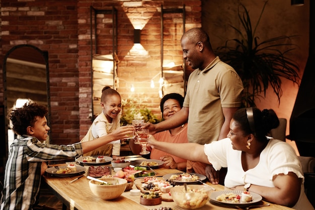 Retrato en tonos cálidos de la feliz familia afroamericana tintineando vasos y brindando mientras disfruta de la cena ...