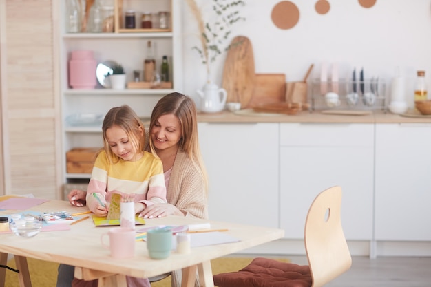 Retrato de tonos cálidos de madre feliz abrazando a su pequeña hija mientras pinta cuadros en la mesa de madera en la cocina, espacio de copia