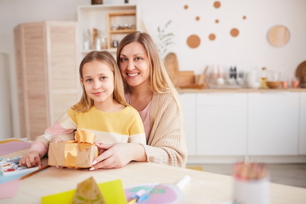 Retrato de tonos cálidos de madre feliz posando con hija mientras abre regalos en la mesa de la cocina de madera, espacio de copia