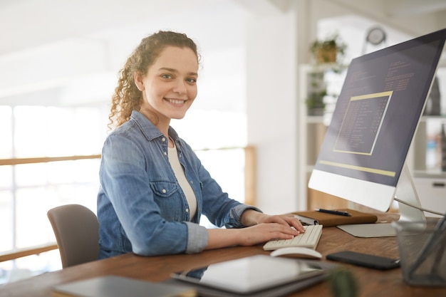 Retrato en tonos cálidos de una mujer joven que usa la computadora y que sonríe a la cámara mientras trabaja en el escritorio en la agencia de desarrollo de software, mujer profesional de TI, espacio de copia