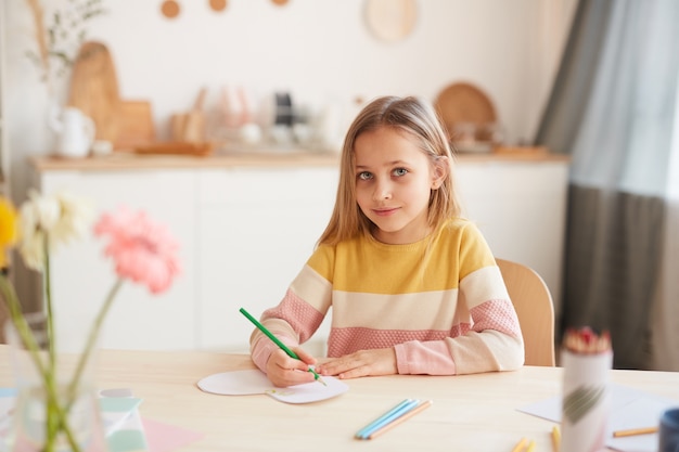 Retrato en tonos cálidos de niña linda sonriendo mientras hace dibujos o hace la tarea mientras está sentado en la mesa en el interior de la casa, espacio de copia