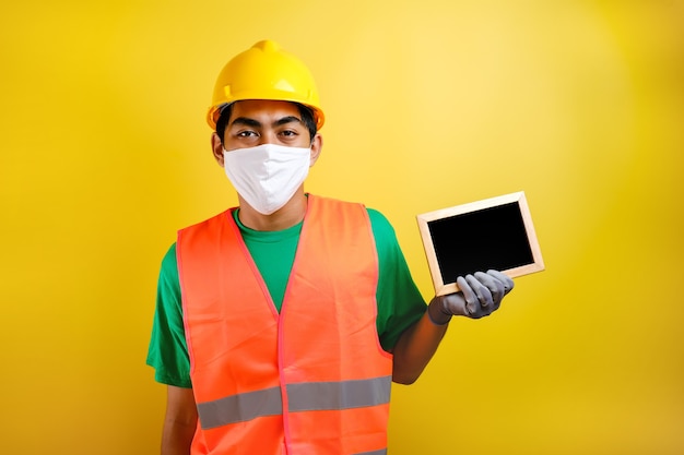 Retrato de trabajador asiático con máscara protectora contra el coronavirus, sosteniendo una pequeña pizarra sobre fondo amarillo