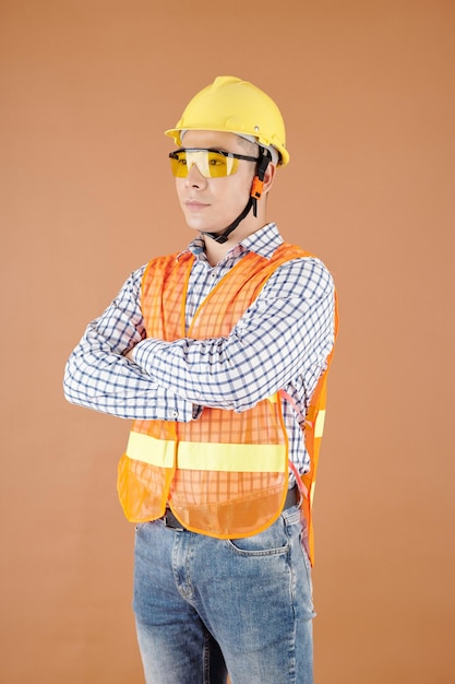 Retrato de un trabajador de la construcción serio cruzando los brazos y mirando hacia otro lado aislado en marrón