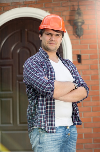 Retrato del trabajador de la construcción sonriente con las manos cruzadas posando contra la puerta de la casa nueva