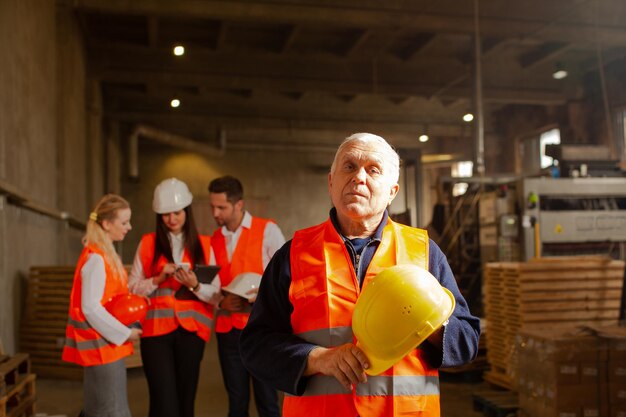 Foto retrato de un trabajador de fábrica experimentado de edad avanzada con su casco en las manos parado en la tienda de una fábrica de madera grupo de jóvenes especialistas en chalecos protectores y cascos parados en el fondo