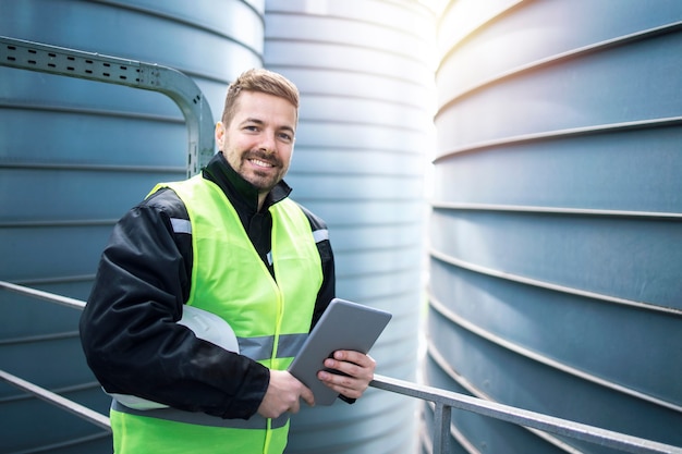 Retrato de trabajador de fábrica con tableta de pie junto a tanques de almacenamiento de silos.