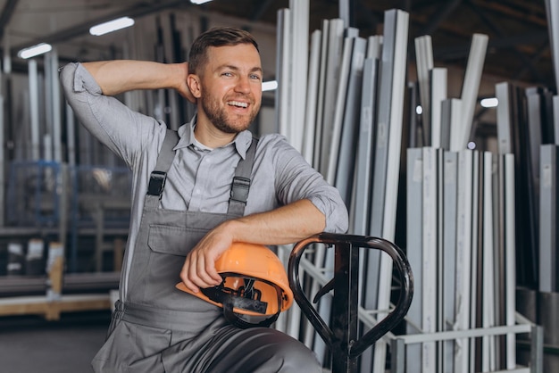 Retrato de un trabajador feliz con un casco naranja y overoles sosteniendo un camión hidráulico contra el fondo de una fábrica y marcos de aluminio