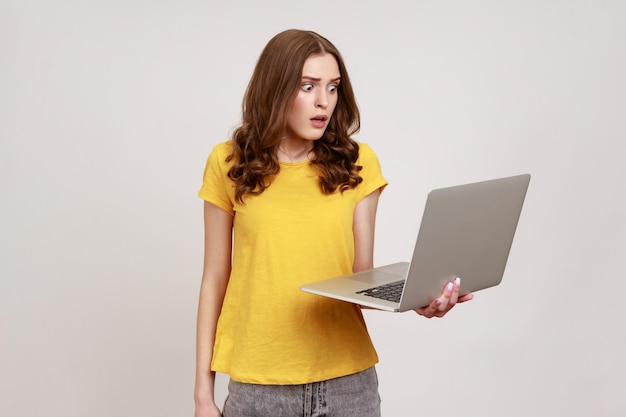 Retrato de un trabajador independiente asustado que trabaja en una computadora Mujer joven sorprendida en una camiseta casual leyendo noticias impactantes en una computadora portátil que trabaja en línea usando una computadora Toma de estudio interior aislada en fondo gris