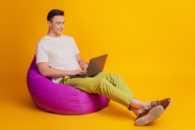 Retrato de un trabajador independiente sentado en una bolsa de frijoles escribiendo en una computadora portátil con una sonrisa dentada en un fondo amarillo