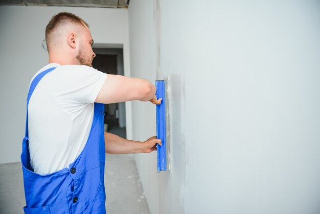 Retrato de un trabajador con un mono y sosteniendo una espátula en sus manos contra el fondo de la pared enlucida. Concepto de trabajo y construcción de reparación