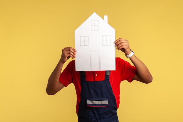 Retrato de un trabajador o un trabajador de mudanzas con un mono azul que cubre la cara con una casa de papel que ofrece ayuda para el servicio de reparación del hogar en la reubicación Foto de estudio interior aislada sobre fondo amarillo