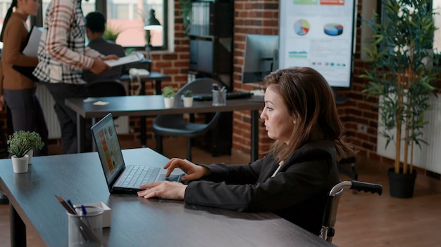 Retrato de un trabajador de oficina en silla de ruedas usando una laptop en el escritorio, creando gráficos de estrategia financiera. Empleado con discapacidad que trabaja en un proyecto empresarial para planificar el desarrollo y el crecimiento.