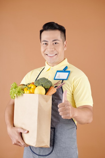 Retrato de un trabajador de supermercado feliz sosteniendo una bolsa de papel con comestibles y mostrando el pulgar hacia arriba