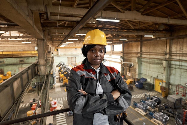 Retrato de una trabajadora africana con casco de trabajo y uniforme parada en el balcón con los brazos cruzados y mirando la cámara