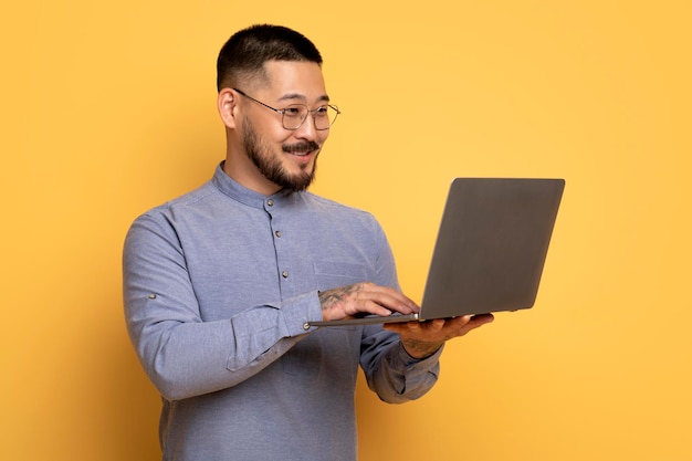 Retrato de trabajo en línea de un hombre asiático sonriente usando una computadora portátil con fondo amarillo