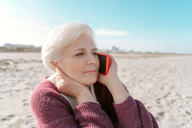 Retrato de una tranquila y atractiva mujer caucásica canosa haciendo una llamada telefónica en la playa