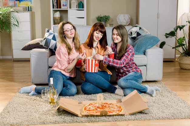 Retrato de tres alegres jóvenes amigas con tazones de palomitas de maíz, pizza y vino, sentados en el suelo cerca del elegante sofá en casa