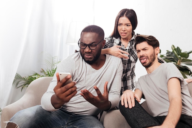 Retrato de tres amigos sentados en sillas en casa y mirando con asombro en el teléfono móvil mientras pasan tiempo juntos aislados