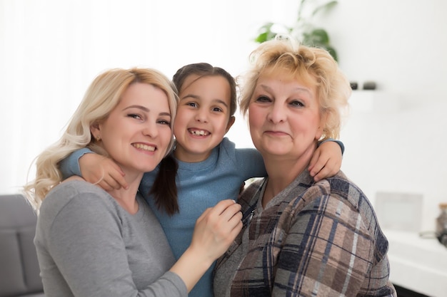 Retrato de tres generaciones de mujeres miran la cámara posando para una foto familiar, linda niña abrazan a mamá y abuela disfrutan del tiempo en casa, sonrientes madre, hija y abuela pasan el fin de semana juntas