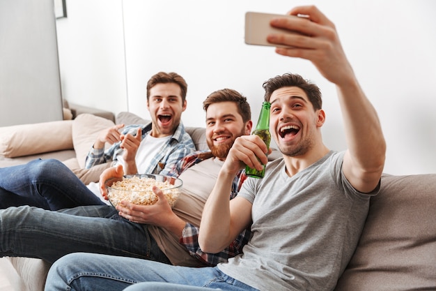 Retrato de tres hombres jóvenes felices tomando un selfie mientras está sentado en casa con cerveza y bocadillos en el interior