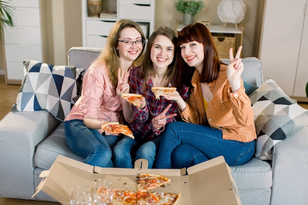 Retrato de tres jóvenes amigas felices en ropa casual comiendo pizza en el sofá en casa. Amistad de mujeres, comer juntas