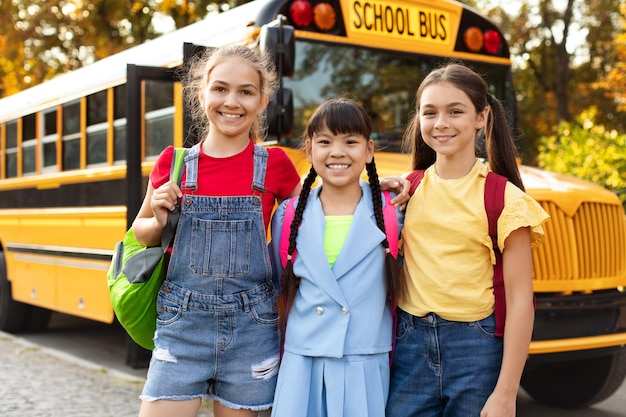 Retrato de tres niñas felices posando juntas al aire libre cerca del autobús escolar