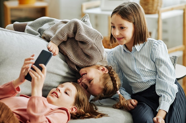 Retrato de tres niñas jugando juntas en el sofá en casa y usando un teléfono inteligente