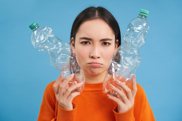 Retrato de una triste mujer asiática con botellas de plástico molesta por la falta de centros de reciclaje de fondo azul.