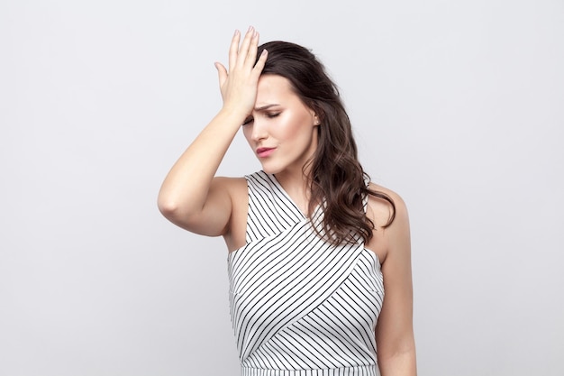 Foto retrato de triste perder hermosa joven morena con maquillaje y vestido a rayas de pie y sosteniendo la mano en la frente. tiro del estudio de interior, aislado en fondo gris.