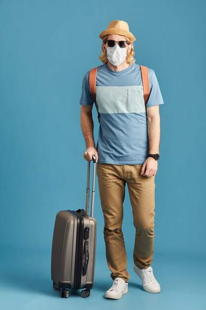 Retrato de turista en máscara y gafas de sol de pie con equipaje y mirando a cámara aislada sobre fondo azul.