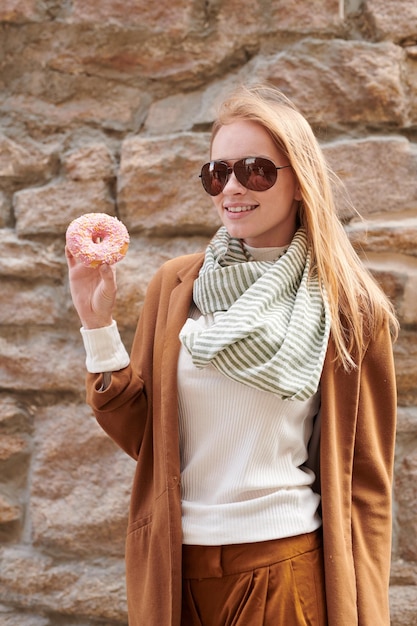 Retrato de una turista positiva y bonita con gafas de sol que muestra una rosquilla con cubierta rosa contra una vieja pared de piedra