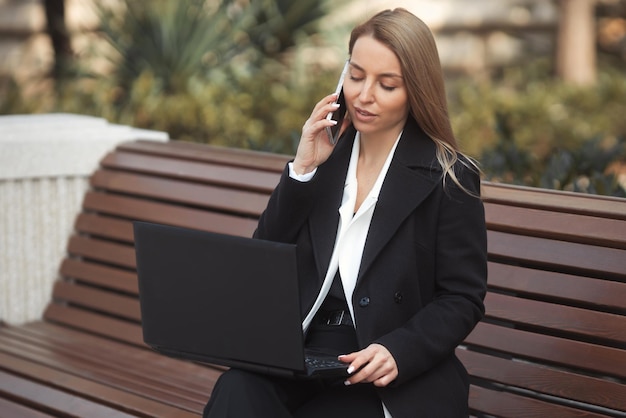 Retrato urbano da bela jovem empresária trabalhando remotamente com laptop e falando ao telefone