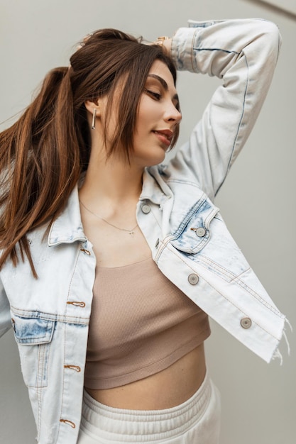 Retrato urbano de jovem morena bonita com penteado na moda jaqueta jeans azul e top poses na rua perto de uma parede cinza