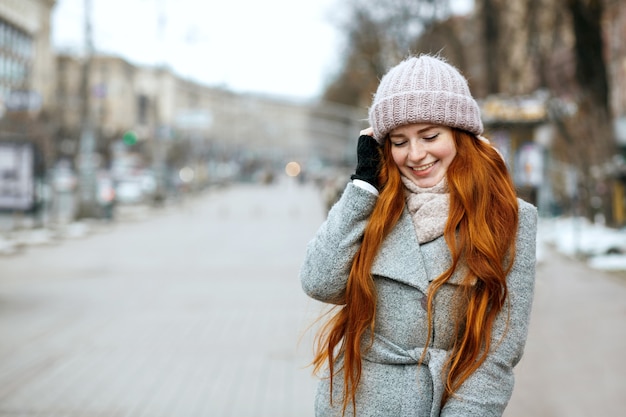 Retrato urbano de feliz niña de jengibre con cabello largo con ropa de abrigo caminando por la ciudad. Espacio para texto